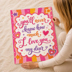 A person sitting on a white carpet holding up a colorful poster that reads You'll never know how much I love you my dear.
