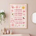 A framed children's affirmation poster hangs on a pink wall above a shelf holding a stuffed bunny, a flower in a vase, and a stack of rings.
