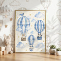 Framed watercolor print showing three blue-striped hot air balloons carrying a puppy, panda, and bunny against a cloudy star background, displayed on a wooden surface with a toy rabbit and potted plants.