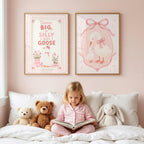 A young girl in pink pajamas sits on a white bed reading a book, with two framed pink nursery art prints hanging on the pink wall above her.