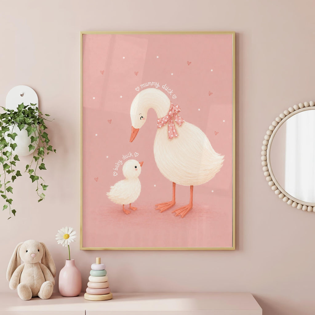 Framed pink nursery art of a mummy duck with a pink bow and a baby duck, hanging on a pink wall next to a plant and round mirror.