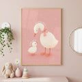Framed pink nursery art of a mummy duck with a pink bow and a baby duck, hanging on a pink wall next to a plant and round mirror.