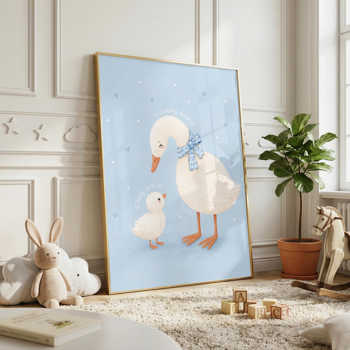 A framed illustration of a mother and baby duck in a gold frame stands on the floor against a white wall.