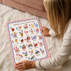 A person with blonde hair sits on a white shag rug, holding the bottom corners of a framed alphabet poster featuring watercolor illustrations.