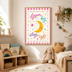A framed nursery art print featuring a smiling moon and the quote dream big sleep tight hangs on a white wall above a low wooden toy shelf in a brightly decorated bedroom.
