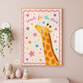 A framed children's print showing a giraffe looking up with the text reach for the stars against a pink star-patterned background, hanging on a pink wall near a white shelf with plants and a toy.