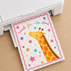 A white home printer feeds out a sheet of paper printed with an illustrated giraffe and the text reach for the stars.