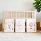 Three framed watercolor prints lean against a light wood crib, featuring pink hot air balloons, clouds, and a personalized name on the center print.