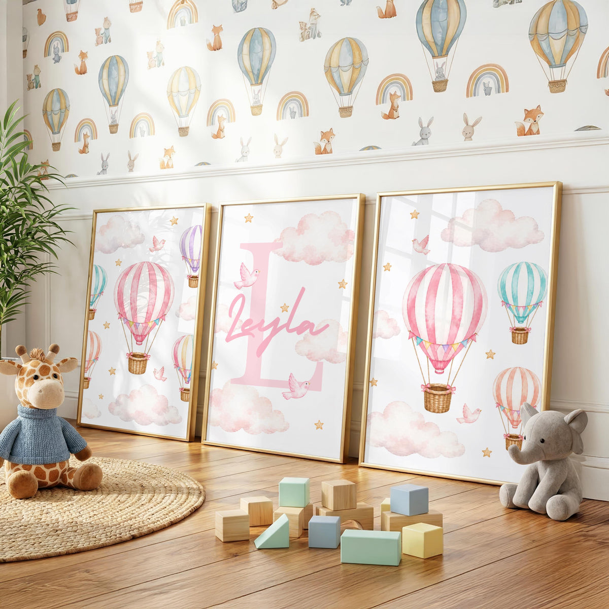 Three framed prints with pink hot air balloons and a personalized name on the center print are displayed on a wooden floor with scattered wooden blocks and plush toys.