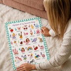 A person in a cream sweater sits on a white shag rug and holds the bottom edges of an illustrated alphabet poster.