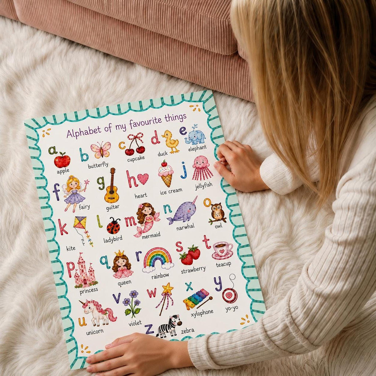 A person in a cream sweater sits on a white shag rug and holds the bottom edges of an illustrated alphabet poster.