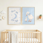 Two framed blue nursery art prints hang above a wooden crib, featuring a moon and stars on the left and two ducks on the right.
