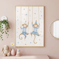 A framed watercolor print of two monkeys on rope swings wearing blue striped outfits and party hats, hanging on a pink wall alongside a stuffed rabbit and wooden toys.