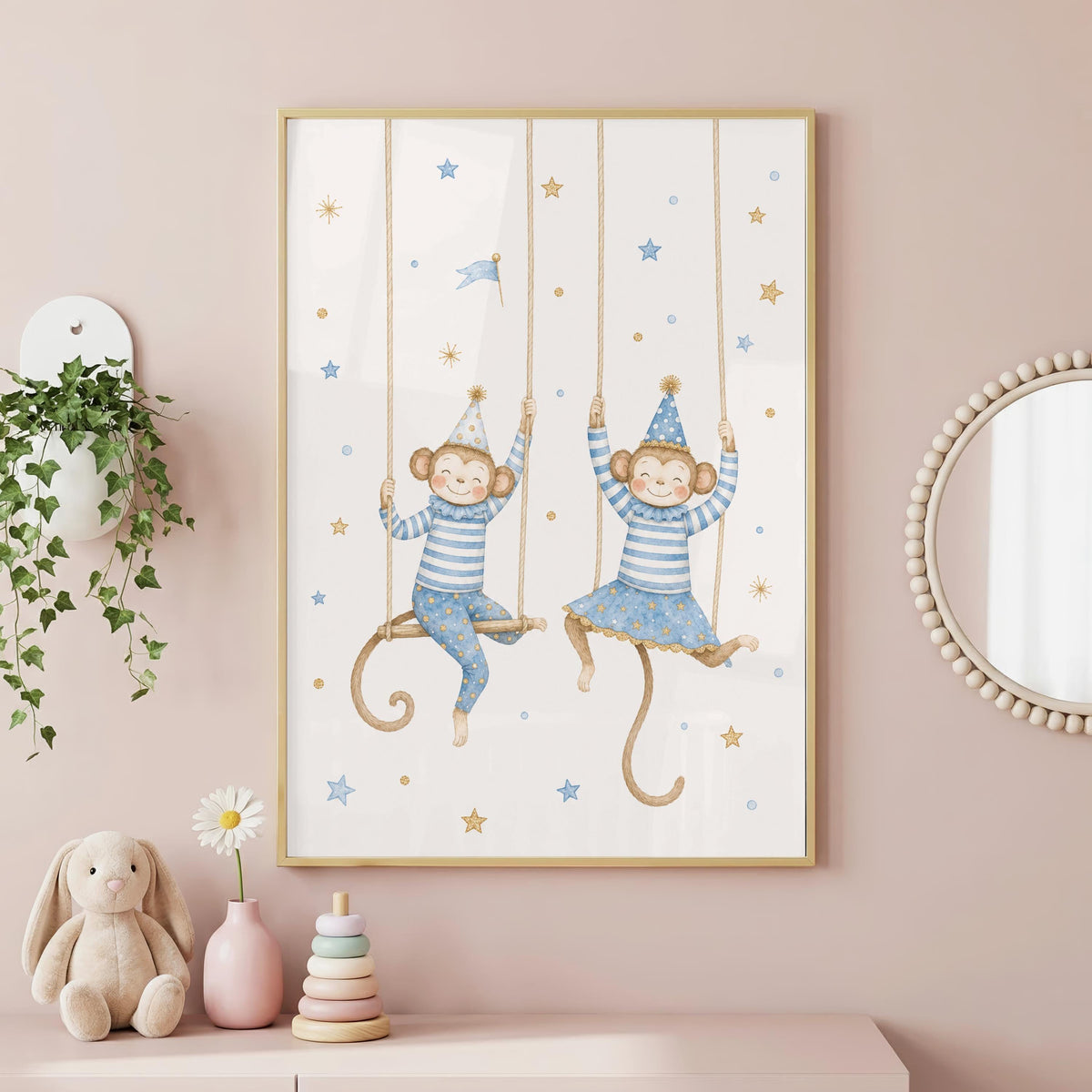 A framed watercolor print of two monkeys on rope swings wearing blue striped outfits and party hats, hanging on a pink wall alongside a stuffed rabbit and wooden toys.