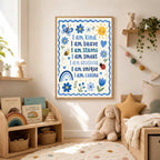 A framed kids' affirmation poster hangs on a white wall next to a wooden shelf holding a plush bear, with a child's desk and toys below.