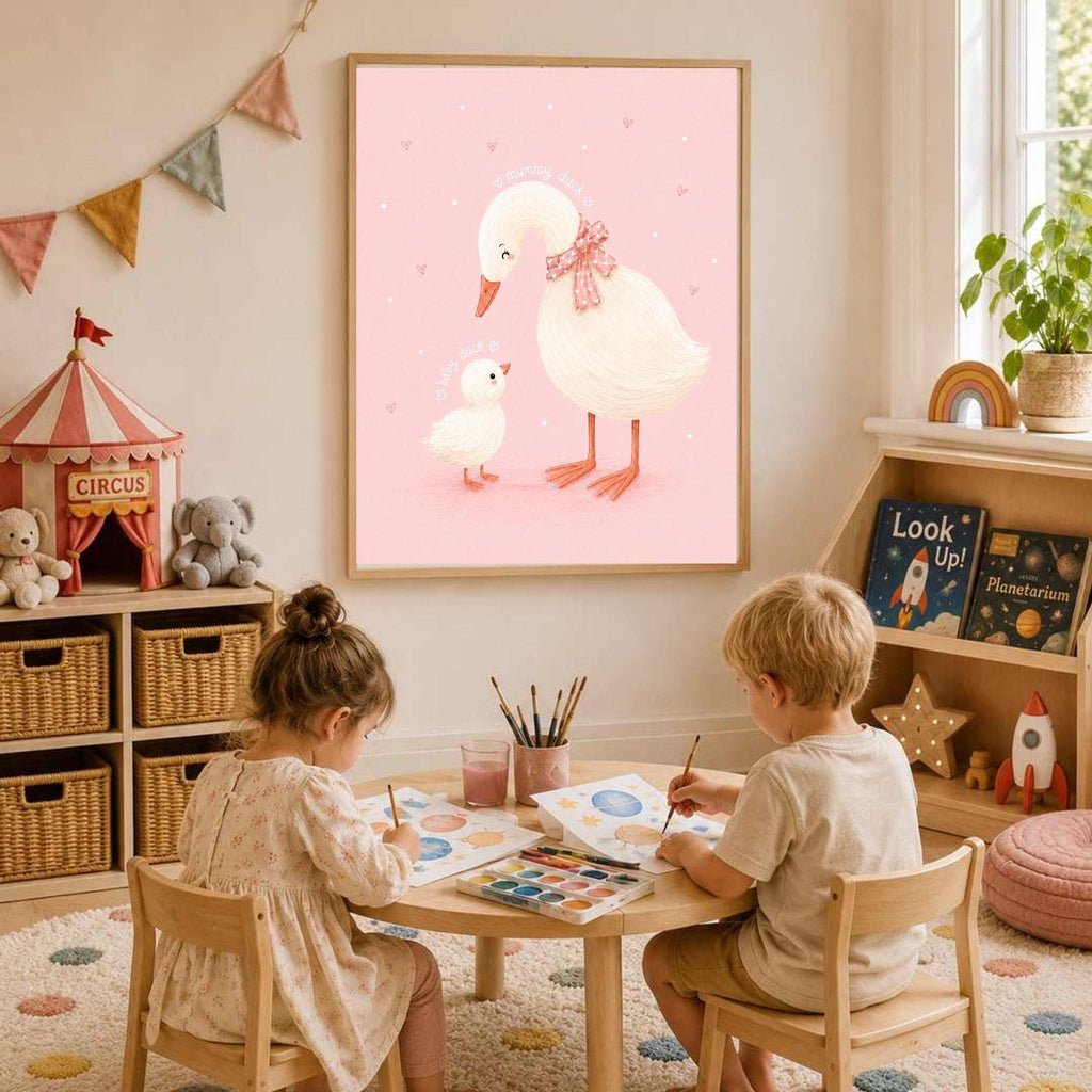Two children sitting at a low wooden table, painting watercolor sheets below a framed pink duck print on the wall.