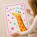 A person sitting on a carpet holds up a large vertical print of an illustrated giraffe surrounded by stars and the text reach for the stars.