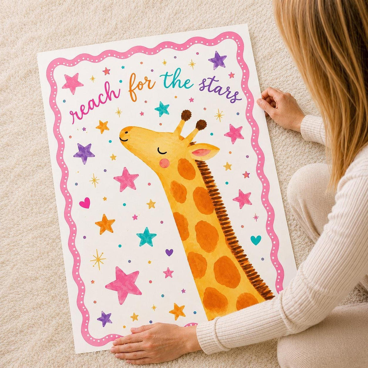A person sitting on a carpet holds up a large vertical print of an illustrated giraffe surrounded by stars and the text reach for the stars.