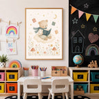 A framed watercolor illustration of a bear reading on a cloud is mounted on a wall above a child's art table and a chalkboard.