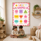 A framed basic shapes poster hangs on a wall above two young children sitting on a rug reading a book.