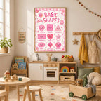 A framed pink educational poster of basic shapes hangs on a white wall above a child play kitchen in a brightly lit playroom.