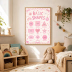 A framed pink Basic Shapes poster is mounted on a white wall above a childrens wooden bookshelf and bed.