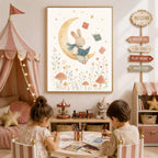 Two children sit at a small table coloring while a framed watercolor illustration of a rabbit reading a book on a crescent moon hangs on the wall above them.